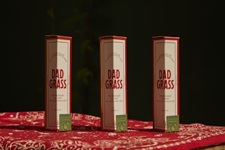 Three packages labeled 'Dad Grass' are placed upright on a surface covered with a red and white patterned cloth. Each package has a smaller, green box with text and a leaf logo on it.