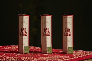 Three packages labeled 'Dad Grass' are placed upright on a surface covered with a red and white patterned cloth. Each package has a smaller, green box with text and a leaf logo on it.