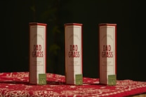 Three packages labeled 'Dad Grass' are placed upright on a surface covered with a red and white patterned cloth. Each package has a smaller, green box with text and a leaf logo on it.