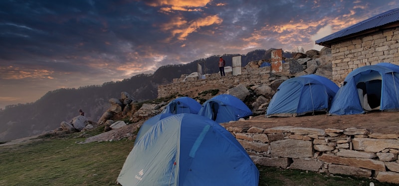 A campsite with several blue tents set up on a grassy area beside a stone structure. The background features a dramatic sky with clouds during sunset, casting an orange hue. There are some people near the tents and rugged terrain with rocks and distant hills.