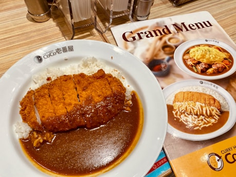 A plate of Japanese curry with crispy breaded cutlet served over white rice. The dish is presented on a white plate with the restaurant logo, accompanied by a menu featuring similar dishes. The table setup includes napkins, salt, and pepper shakers.