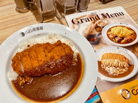 A plate of Japanese curry with crispy breaded cutlet served over white rice. The dish is presented on a white plate with the restaurant logo, accompanied by a menu featuring similar dishes. The table setup includes napkins, salt, and pepper shakers.