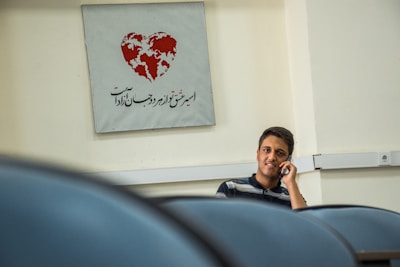 A young man is sitting and talking on a phone in a room with cream-colored walls. Behind him, there is a sign featuring a heart-shaped design with a map inside it and some text in Persian. Part of the furniture or seating arrangement appears in the foreground.