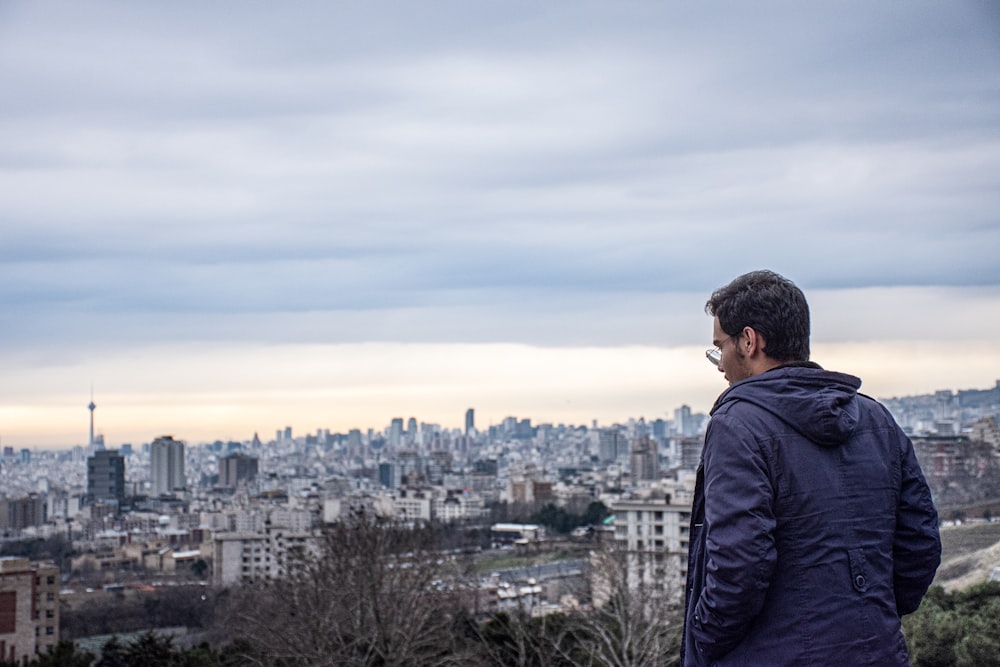 검은 재킷을 입은 남자가 낮에 도시를 바라보는 건물 꼭대기에 서 있다