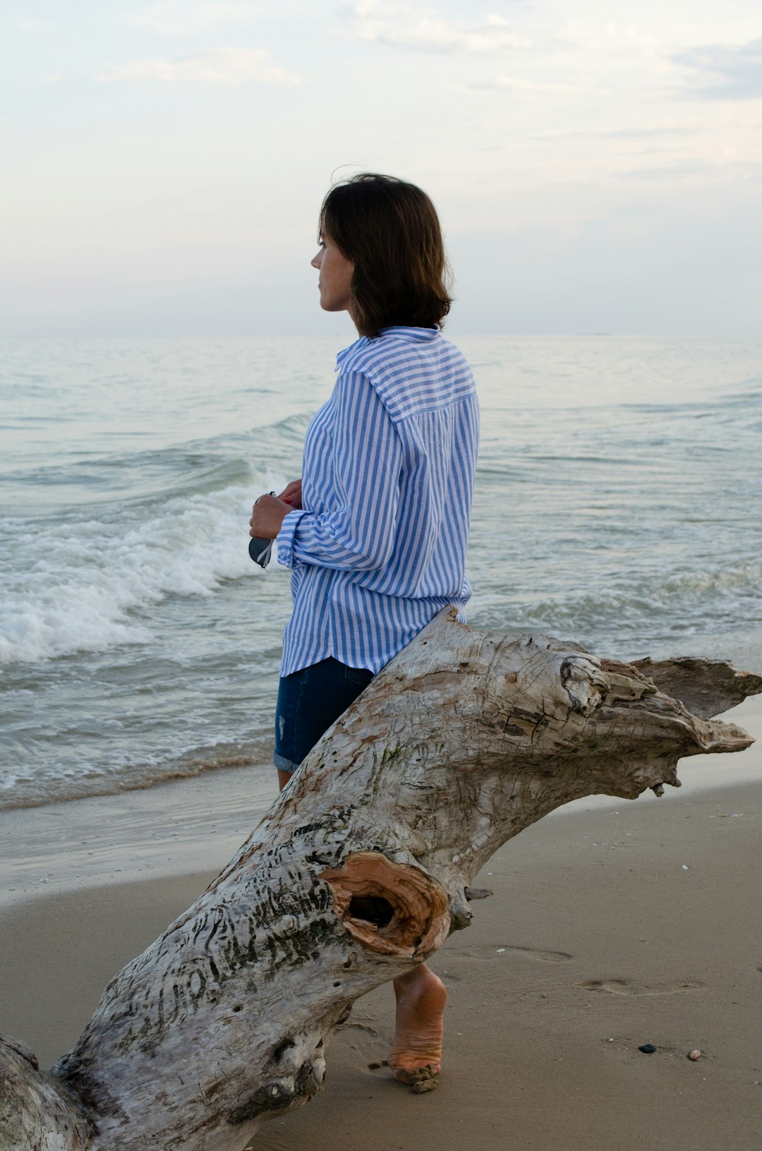 woman in white and blue stripe shirt sitting on brown tree trunk on beach during daytime