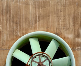 A large industrial ventilation fan with pale green blades set against a textured, rust-colored wall. The fan has a protective grid in front of it, and the wall shows vertical streaks, possibly from weathering.