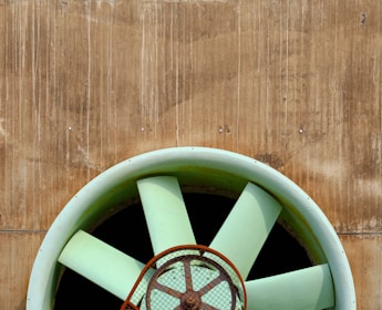 A large industrial ventilation fan with pale green blades set against a textured, rust-colored wall. The fan has a protective grid in front of it, and the wall shows vertical streaks, possibly from weathering.