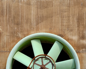 A large industrial ventilation fan with pale green blades set against a textured, rust-colored wall. The fan has a protective grid in front of it, and the wall shows vertical streaks, possibly from weathering.