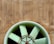 A large industrial ventilation fan with pale green blades set against a textured, rust-colored wall. The fan has a protective grid in front of it, and the wall shows vertical streaks, possibly from weathering.