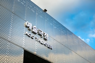 A modern building facade with a perforated metal panel design, featuring a sign with Korean characters and the words 'ACC THEATER' in English. The sky in the background is blue with some clouds.