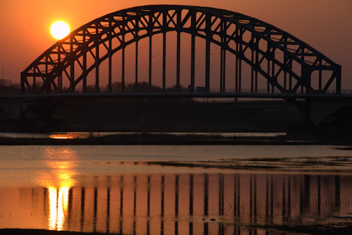 A sleek steel bridge arching gracefully over a calm river at sunset.