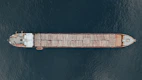 Aerial view of a large cargo ship transporting logs across a body of water. The vessel is long and narrow, with logs neatly stacked along its length. The ship contrasts against the deep blue water surrounding it.