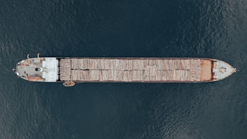 Aerial view of a large cargo ship transporting logs across a body of water. The vessel is long and narrow, with logs neatly stacked along its length. The ship contrasts against the deep blue water surrounding it.