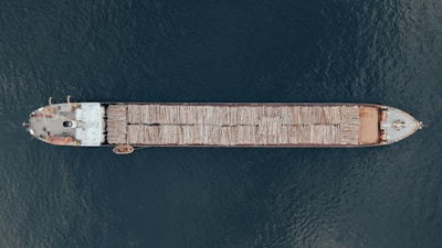 Aerial view of a large cargo ship transporting logs across a body of water. The vessel is long and narrow, with logs neatly stacked along its length. The ship contrasts against the deep blue water surrounding it.