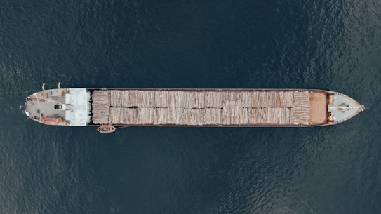 Aerial view of a large cargo ship transporting logs across a body of water. The vessel is long and narrow, with logs neatly stacked along its length. The ship contrasts against the deep blue water surrounding it.