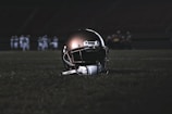 A football helmet rests on the grass of a dimly lit football field. In the background, football players appear in a huddle, dressed in uniforms and gear, slightly out of focus.
