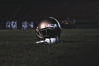 A football helmet rests on the grass of a dimly lit football field. In the background, football players appear in a huddle, dressed in uniforms and gear, slightly out of focus.