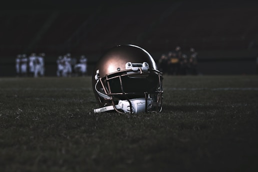 A football helmet rests on the grass of a dimly lit football field. In the background, football players appear in a huddle, dressed in uniforms and gear, slightly out of focus.