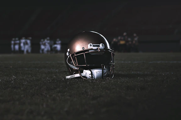 A dynamic image showing an American football helmet resting on a black and gold field under bright stadium lights.
