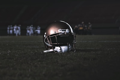 A football helmet rests on the grass of a dimly lit football field. In the background, football players appear in a huddle, dressed in uniforms and gear, slightly out of focus.