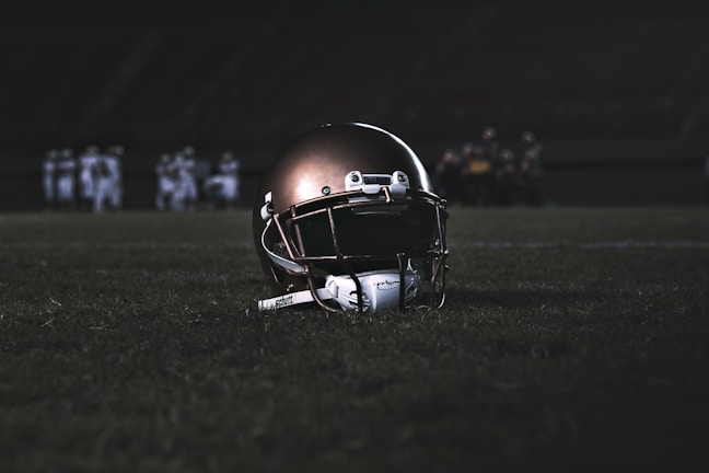A football helmet rests on the grass of a dimly lit football field. In the background, football players appear in a huddle, dressed in uniforms and gear, slightly out of focus.