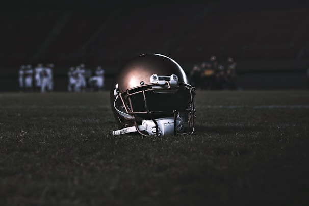 A football helmet rests on the grass of a dimly lit football field. In the background, football players appear in a huddle, dressed in uniforms and gear, slightly out of focus.