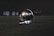 A football helmet rests on the grass of a dimly lit football field. In the background, football players appear in a huddle, dressed in uniforms and gear, slightly out of focus.