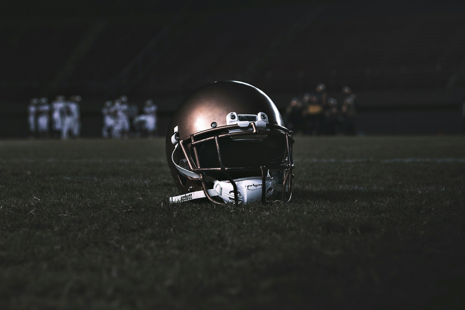 A football helmet rests on the grass of a dimly lit football field. In the background, football players appear in a huddle, dressed in uniforms and gear, slightly out of focus.