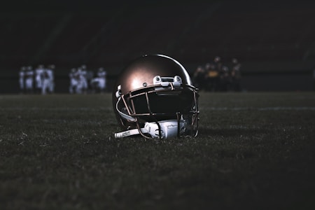 A football helmet rests on the grass of a dimly lit football field. In the background, football players appear in a huddle, dressed in uniforms and gear, slightly out of focus.