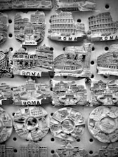 A collection of intricately designed fridge magnets representing famous landmarks in Rome, displayed on a perforated board. Details include well-known architecture such as the Colosseum and other historical structures, with the word 'ROMA' prominently featured.