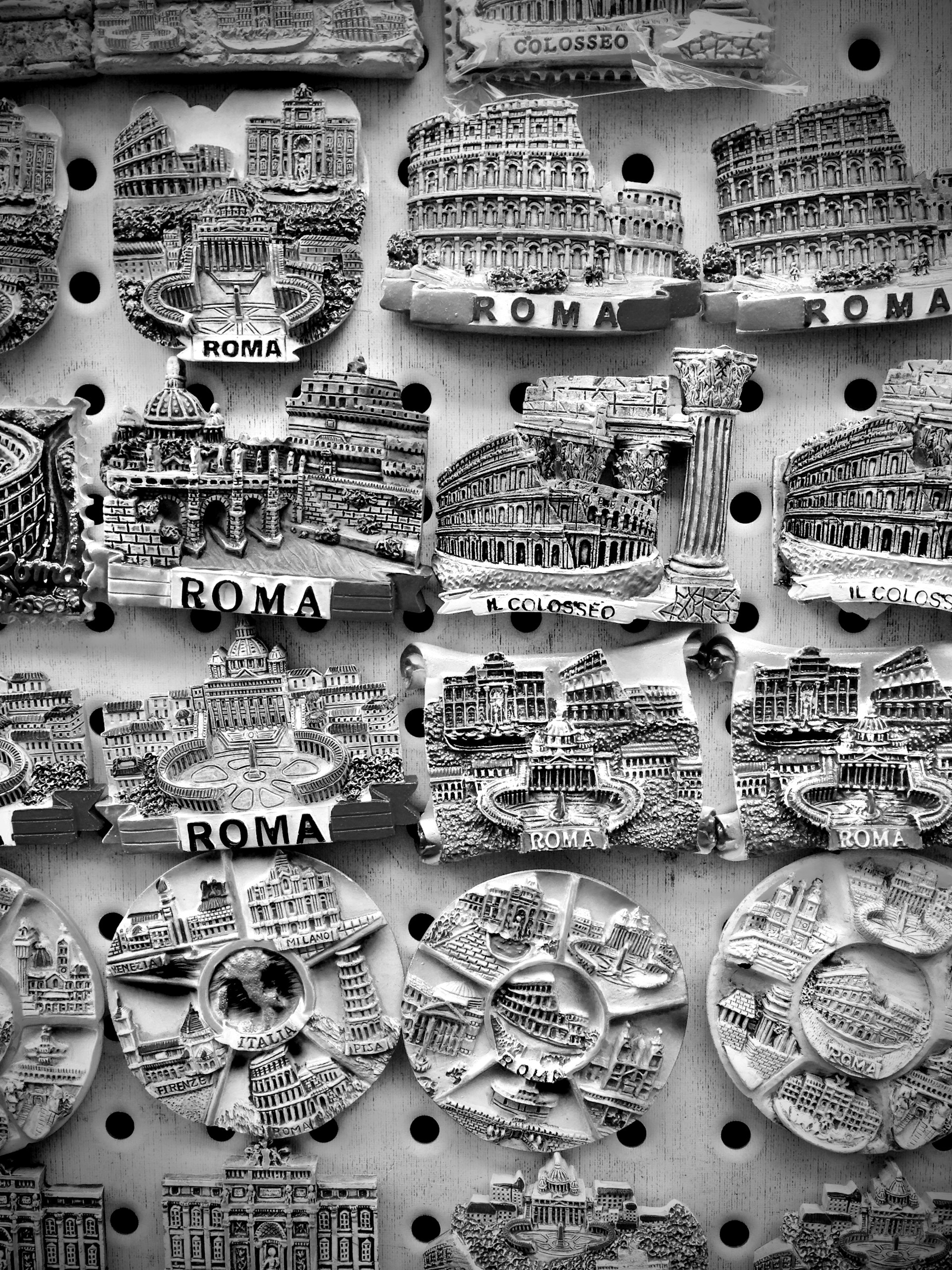 A collection of intricately designed fridge magnets representing famous landmarks in Rome, displayed on a perforated board. Details include well-known architecture such as the Colosseum and other historical structures, with the word 'ROMA' prominently featured.