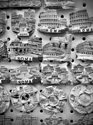 A collection of intricately designed fridge magnets representing famous landmarks in Rome, displayed on a perforated board. Details include well-known architecture such as the Colosseum and other historical structures, with the word 'ROMA' prominently featured.