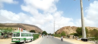 A rural road scene with a small green and white bus parked on the left side. Motorcyclists are riding on the road in the midst of a clear day with scattered clouds. Power lines run alongside the road, and there are hills and greenery in the background.