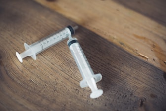 Two plastic syringes are placed on a wooden surface with measurements visible, indicating their use for precise liquid administration.