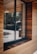 A modern building exterior features a large window set within wooden paneling. The wood panels are arranged horizontally, showcasing a natural brown hue. Reflections of trees and a road can be seen in the glass, along with a pendant light hanging from the ceiling inside.