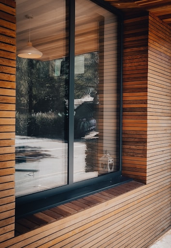 A modern building exterior features a large window set within wooden paneling. The wood panels are arranged horizontally, showcasing a natural brown hue. Reflections of trees and a road can be seen in the glass, along with a pendant light hanging from the ceiling inside.