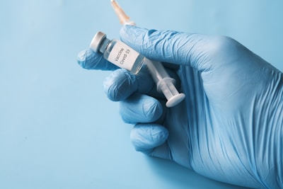 A gloved hand is holding a syringe and a small vial labeled 'Covid-19 Vaccine' against a light blue background.