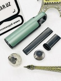 A collection of objects is displayed on a white textured surface. Among the items is a green and black cylindrical device, an attachment nozzle, a small brush, a transparent container, and a metal filter. In the background, there are parts of a model resembling a metallic Eiffel Tower and a white and black sign with text.