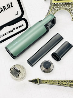 A collection of objects is displayed on a white textured surface. Among the items is a green and black cylindrical device, an attachment nozzle, a small brush, a transparent container, and a metal filter. In the background, there are parts of a model resembling a metallic Eiffel Tower and a white and black sign with text.