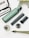 A collection of objects is displayed on a white textured surface. Among the items is a green and black cylindrical device, an attachment nozzle, a small brush, a transparent container, and a metal filter. In the background, there are parts of a model resembling a metallic Eiffel Tower and a white and black sign with text.