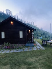 A secluded wooden cabin with lit lamps is surrounded by lush greenery and colorful flower beds. It sits on a patch of grass with a stone pathway leading to the entrance. In the background, rolling hills covered in dense trees are visible under an overcast sky. On the right, there is an outdoor seating area with a table and chairs.