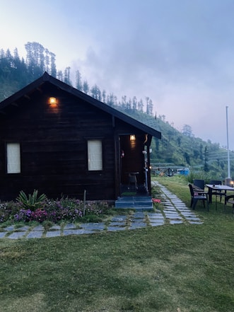 A secluded wooden cabin with lit lamps is surrounded by lush greenery and colorful flower beds. It sits on a patch of grass with a stone pathway leading to the entrance. In the background, rolling hills covered in dense trees are visible under an overcast sky. On the right, there is an outdoor seating area with a table and chairs.