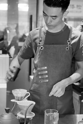 A person is wearing an apron and is engaged in the process of making coffee using a pour-over method. The scene includes coffee drippers and a kettle on a table, indicating a manual brewing setup.
