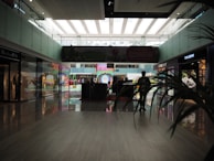 A modern indoor shopping mall with high ceilings and glass walls. The area is lit with natural light coming from the large windows above. There are various storefronts including a T.M. Lewin and Tag Heuer. A few people are walking or standing around, and there are some decorative elements such as arches and bright colors visible in the background. A large digital advertisement panel is centrally located.