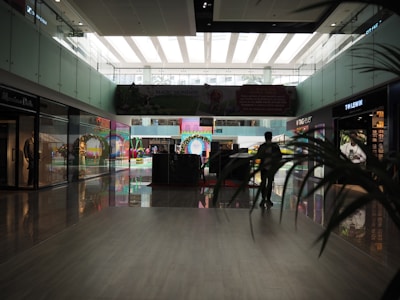 A modern indoor shopping mall with high ceilings and glass walls. The area is lit with natural light coming from the large windows above. There are various storefronts including a T.M. Lewin and Tag Heuer. A few people are walking or standing around, and there are some decorative elements such as arches and bright colors visible in the background. A large digital advertisement panel is centrally located.