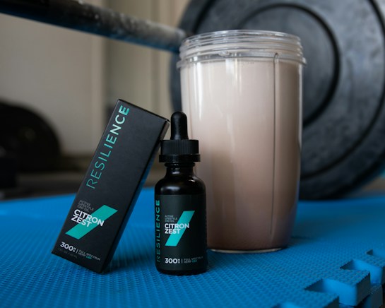 A bottle of Resilience CBD oil, labeled 'Citron Zest', is positioned next to its packaging box on a blue exercise mat. Beside it, there is a shaker filled with a light-colored drink. In the background, there is a blurred fitness weight, suggesting a workout setting.