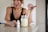 A woman with earrings and wearing a black top is smiling while using a dropper to add liquid from a small white bottle labeled 'resilience' into a jar filled with a light green smoothie. The woman is standing by a countertop in a brightly lit kitchen.