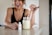 A woman with earrings and wearing a black top is smiling while using a dropper to add liquid from a small white bottle labeled 'resilience' into a jar filled with a light green smoothie. The woman is standing by a countertop in a brightly lit kitchen.