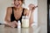 A woman with earrings and wearing a black top is smiling while using a dropper to add liquid from a small white bottle labeled 'resilience' into a jar filled with a light green smoothie. The woman is standing by a countertop in a brightly lit kitchen.
