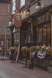 The image depicts a historical tavern with a vintage look on a brick street. The building has a sign reading 'Green Dragon Tavern' and is adorned with American decorations, including flags and banners. There are flower boxes with vibrant flowers under the windows and an umbrella with the Guinness logo. A mannequin dressed in colonial-era military attire stands near the entrance.
